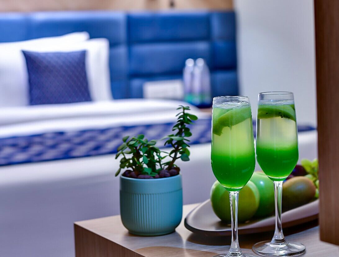 Habitación de hotel con una cama, una mesita de noche con una planta en maceta y dos vasos de bebida verde.