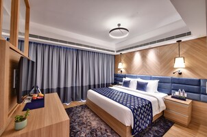 A modern hotel room with a large bed, a TV, a desk with a plant, and a nightstand with a lamp.
