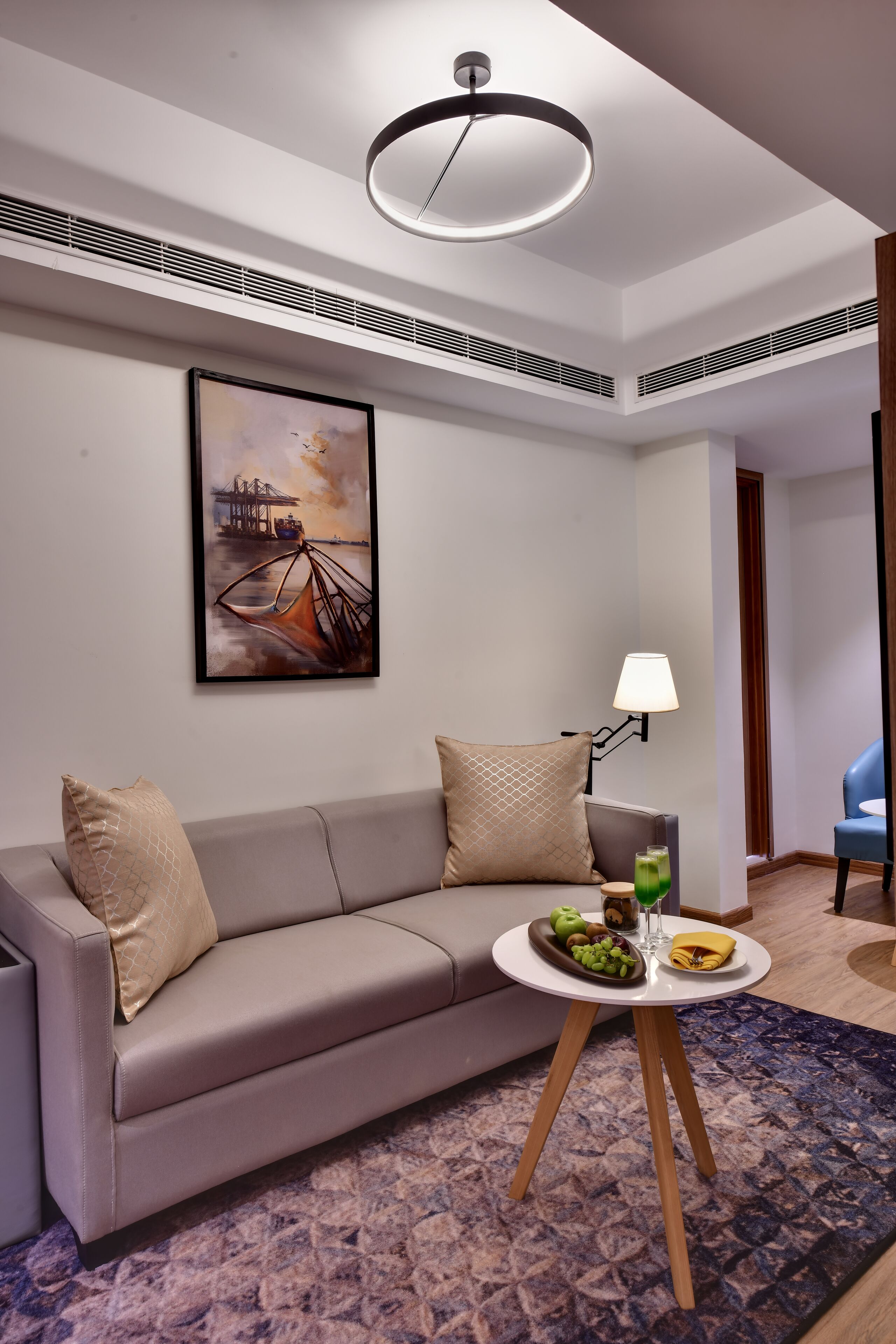 A modern living room with a grey sofa, a round coffee table with fruits and a glass, a painting on the wall, and a blue chair in the background.