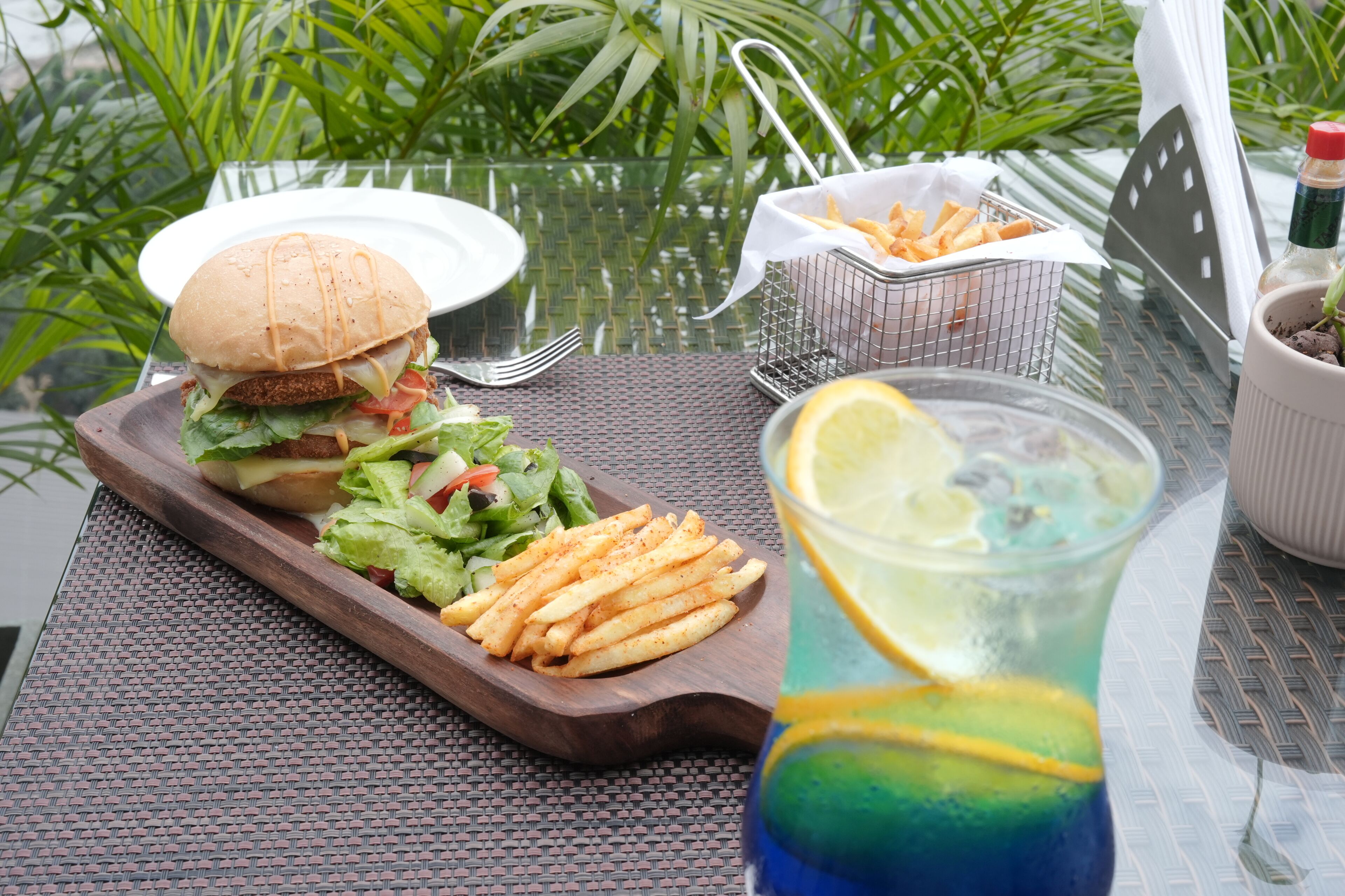 A meal with a burger, fries, and a drink on a table with a plant in the background.