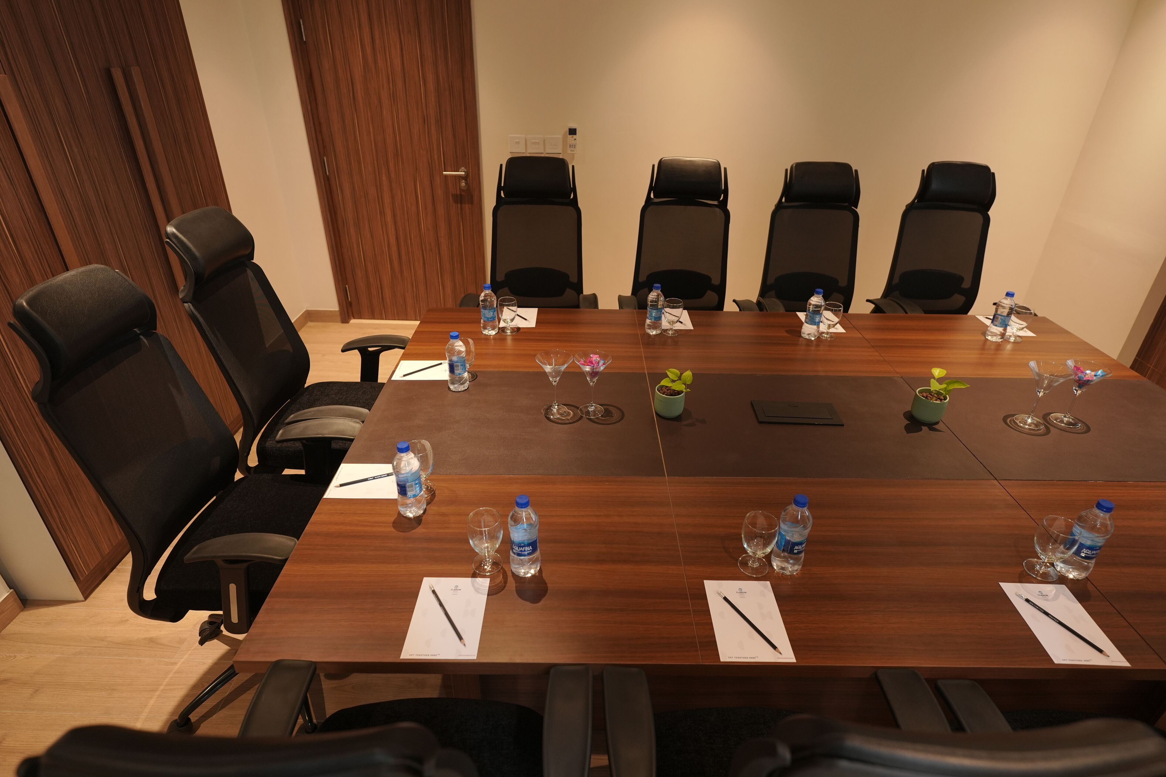 Un salón de conferencias con una larga mesa, seis sillas negras, botellas de agua, vasos y una planta pequeña.