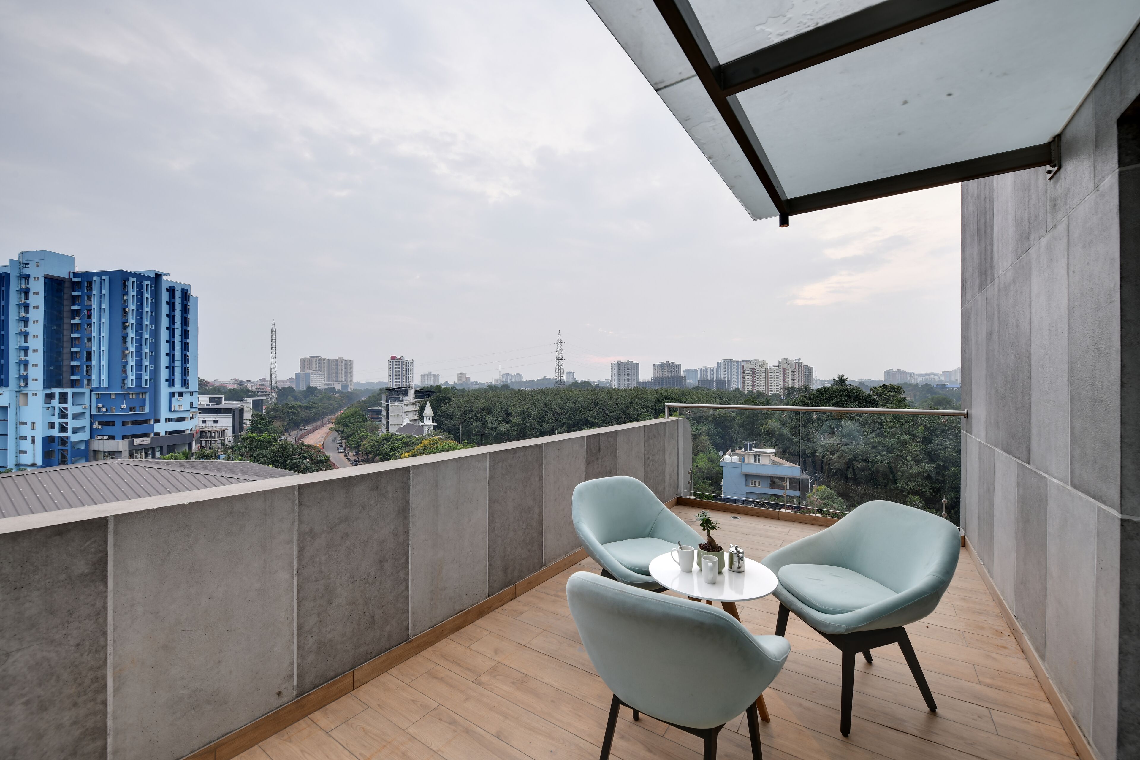 A balcony with two light blue chairs and a small table, overlooking a cityscape with high-rise buildings and greenery.