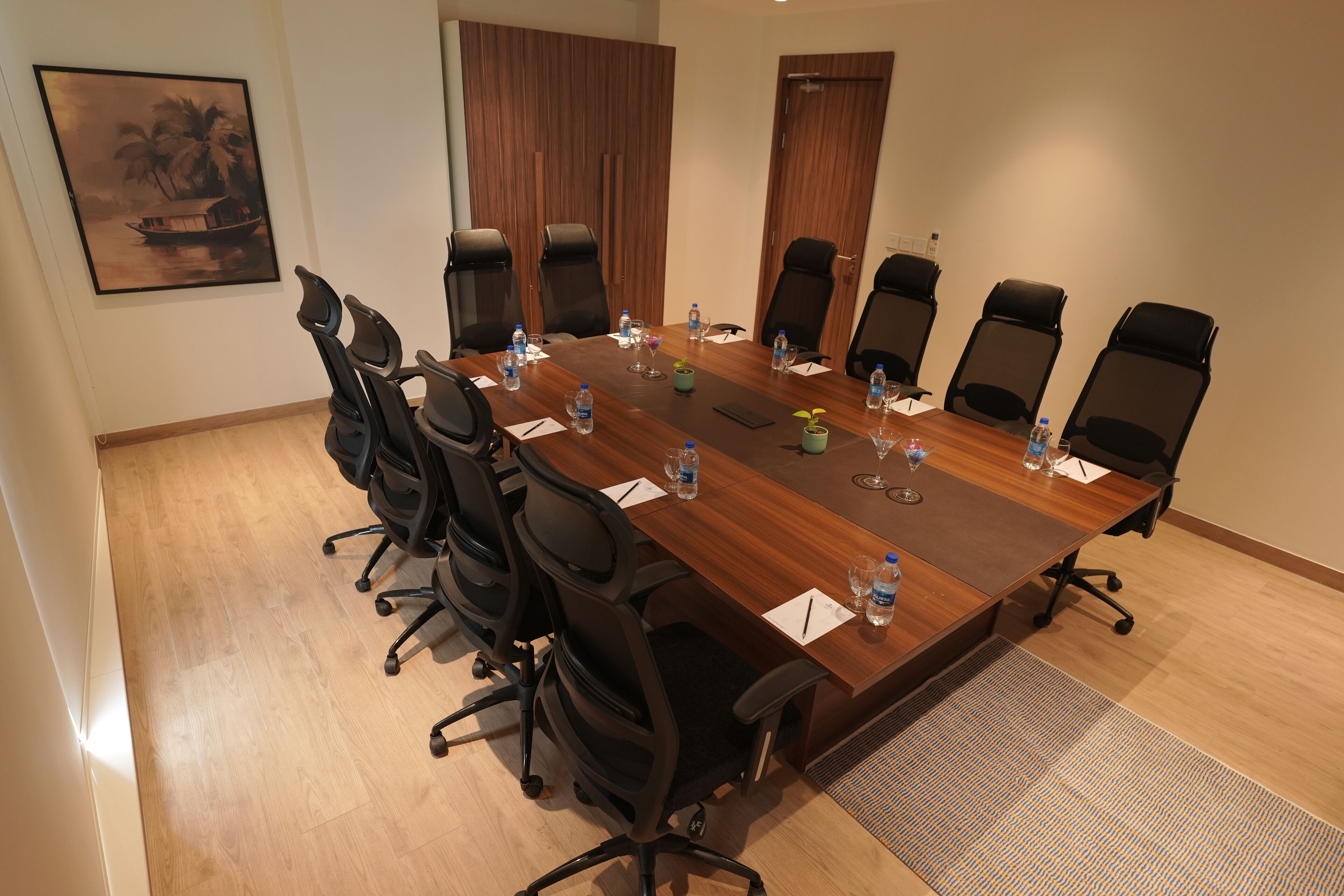 Une salle de conférence avec une table en bois, des chaises de bureau noires, des bouteilles d’eau et un tableau encadré accroché au mur.