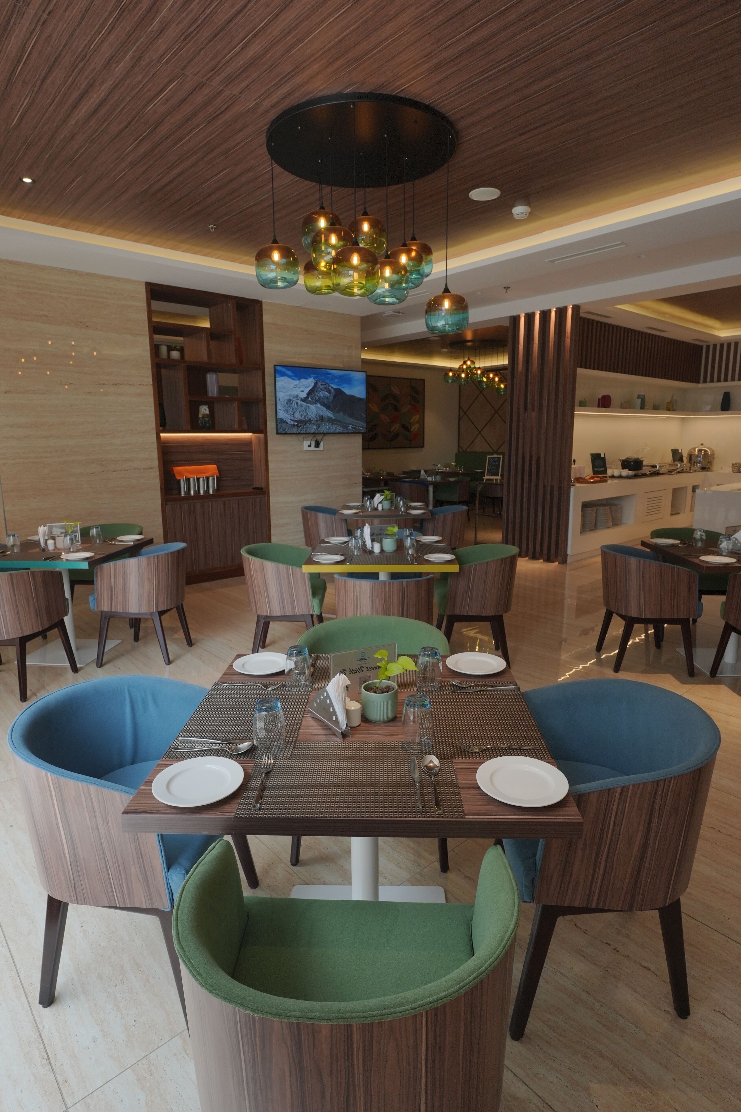 A modern dining area with wooden tables and green upholstered chairs, a central chandelier, and a television mounted on the wall.
