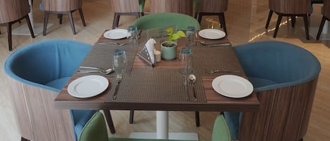 A modern dining area with wooden tables and green upholstered chairs, a central chandelier, and a television mounted on the wall.