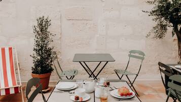 Une table dressée pour un repas, avec des assiettes, des tasses et une théière, entourée de chaises et de plantes en pot.