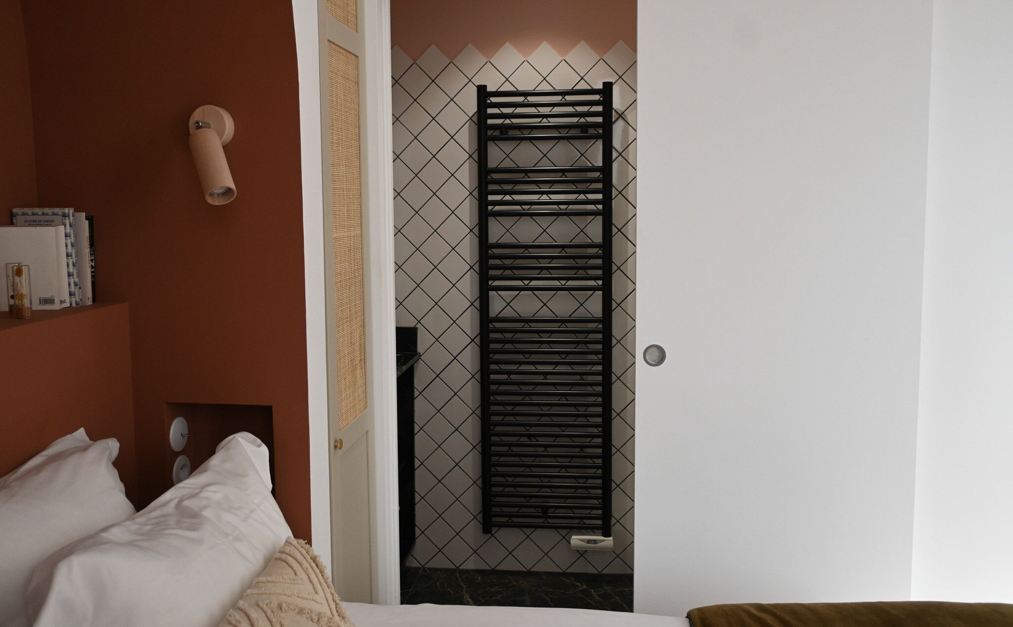 Habitación de hotel con una cama, una mesita de noche con libros, un perchero de pared y una puerta con un papel tapiz de diseño geométrico.