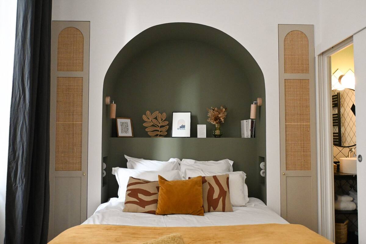 Um quarto com uma grande cabeceira arqueada, cama com roupa de cama branca e almofadas estampadas, prateleira fixada na parede com objetos decorativos e uma porta que dá para o banheiro.