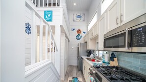 Une cuisine étroite avec des meubles blancs, un four à micro-ondes et une cuisinière à gaz. Un tapis décoratif orné d’un motif de sirène recouvre le sol.