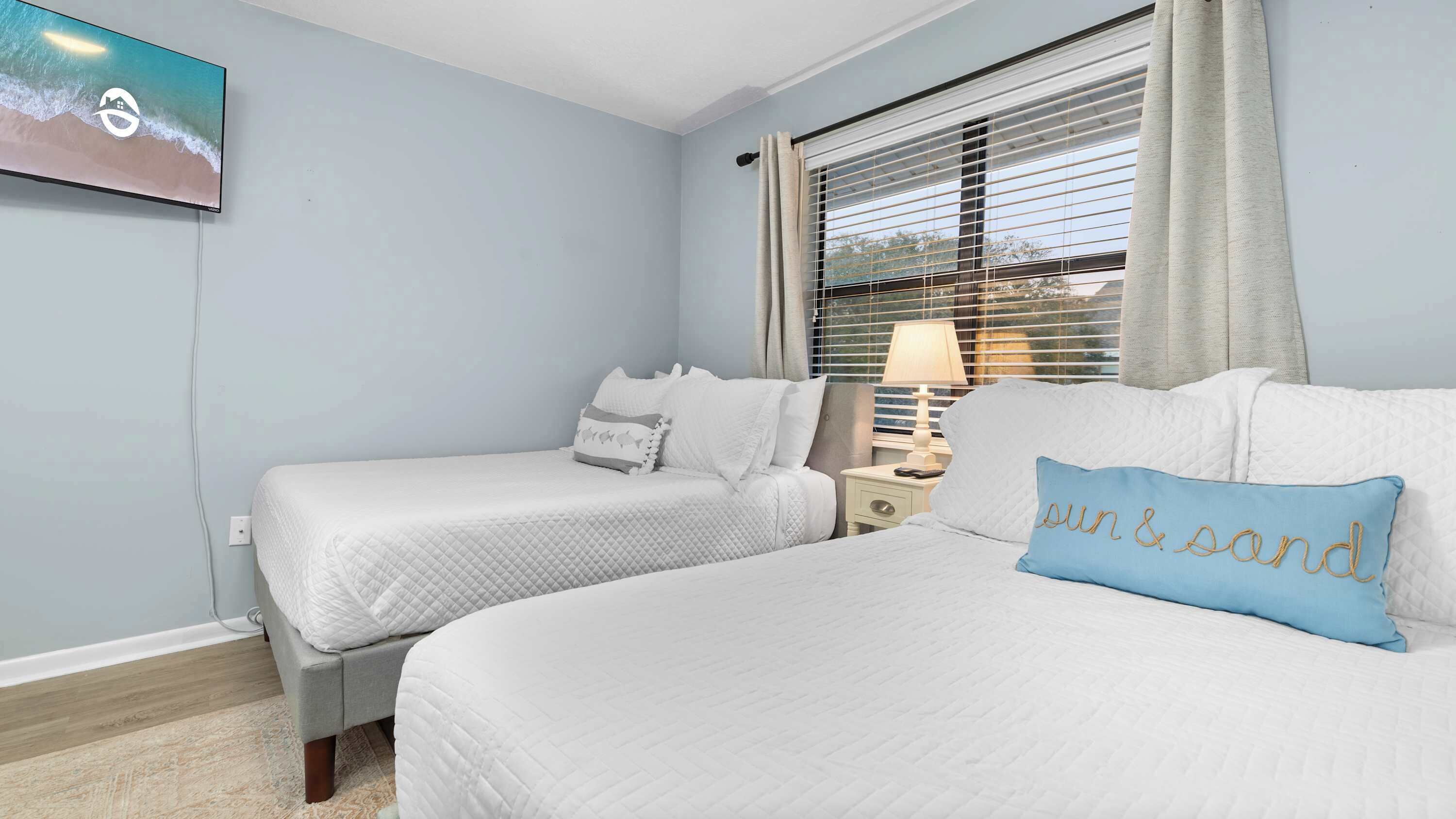 A hotel room with two beds, a television on the wall, a window with blinds, and a nightstand with a lamp.