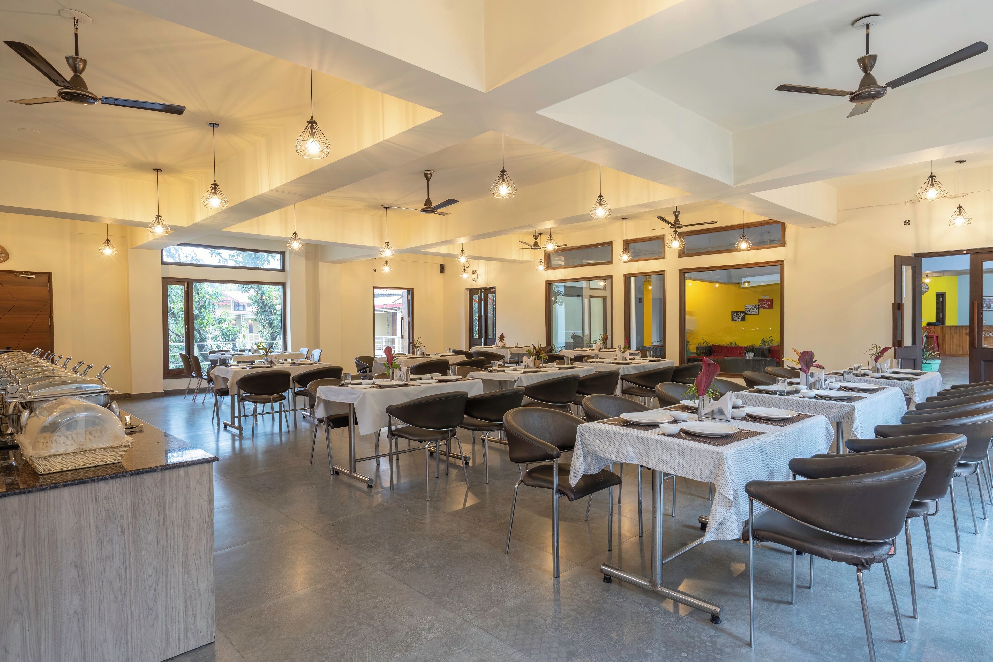 A spacious dining area with multiple tables and chairs, ceiling fans, and natural light from large windows.