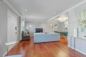 A spacious living room with a large grey sofa, a small table, and a dining area with green chairs.