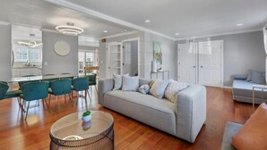 A modern living room with a grey sofa, teal chairs, a round coffee table, and a dining area with a white table and chairs. - @ Marbella Lane - Buckingham Place Laundry + P (Pacifica)