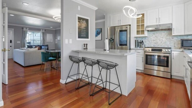 A modern kitchen with white cabinets, stainless steel appliances, and a bar area with stools. - @ Marbella Lane - Buckingham Place Laundry + P (Pacifica)