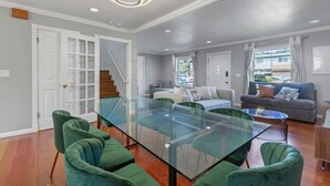 A modern living room with a glass-top dining table, green upholstered chairs, a grey sofa, and a staircase leading to another level. - @ Marbella Lane - Buckingham Place Laundry + P (Pacifica)