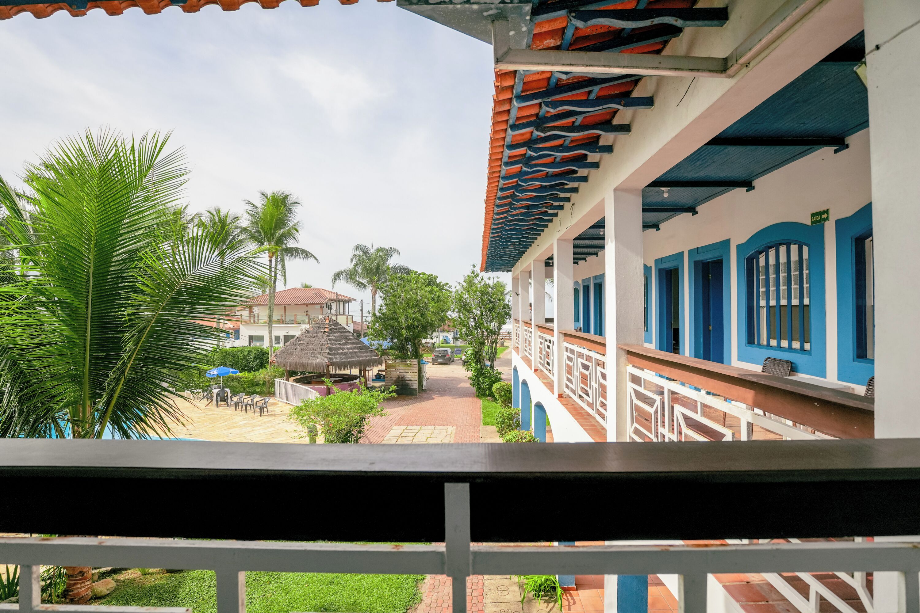 A view from a balcony overlooking a courtyard with palm trees, a thatched-roof structure, and a building with blue and white walls and a tiled roof.
