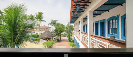 A view from a balcony overlooking a courtyard with palm trees, a thatched-roof structure, and a building with blue and white walls and a tiled roof.