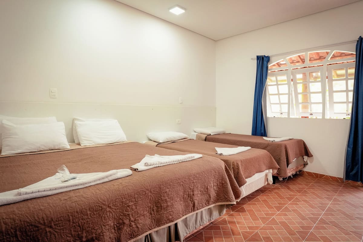 Habitación de hotel con dos camas, cada una con una colcha marrón y almohadas blancas. Hay una ventana con cortinas azules y vistas al exterior.