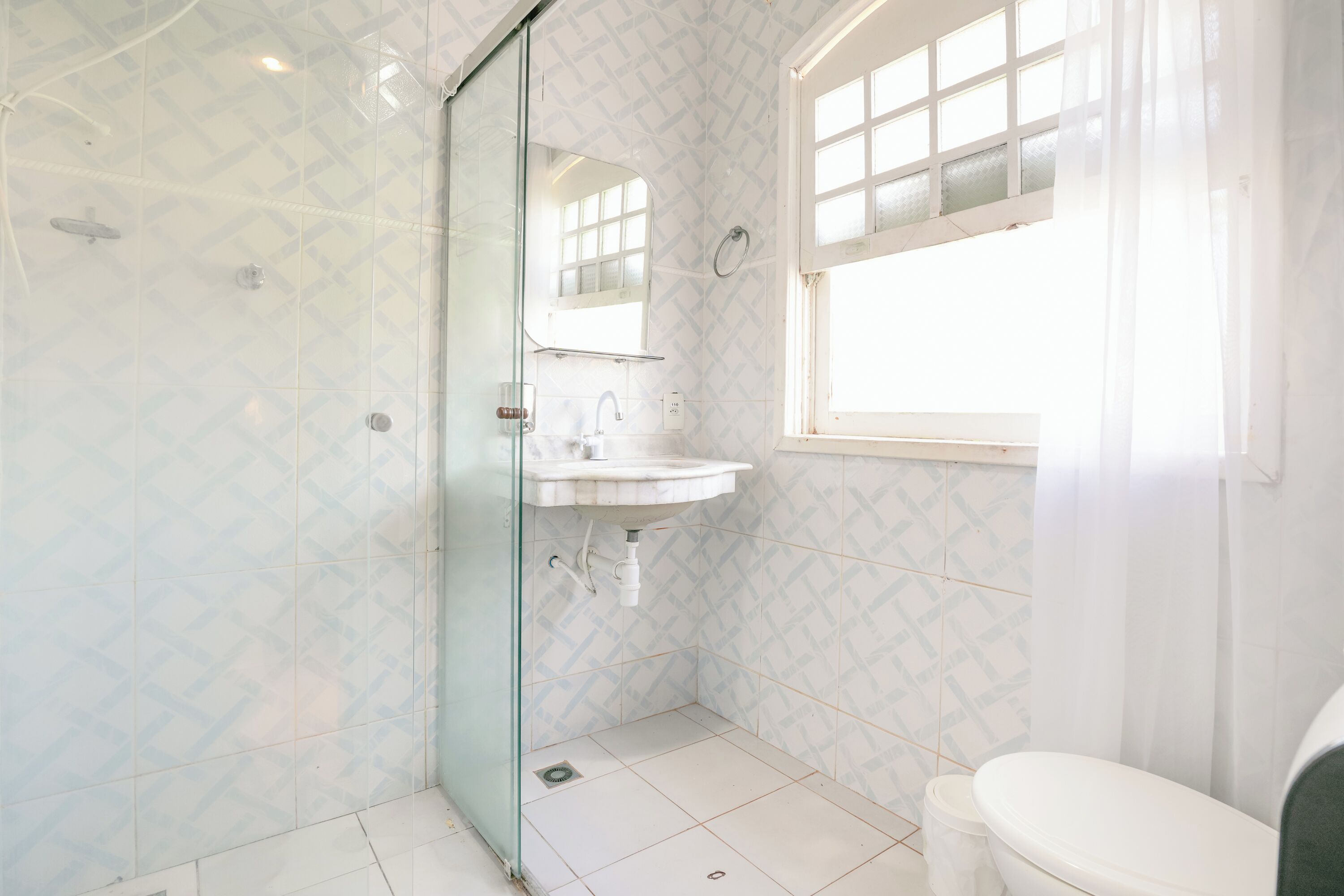 A bathroom with a glass shower door, a sink, a toilet, and a window with curtains.