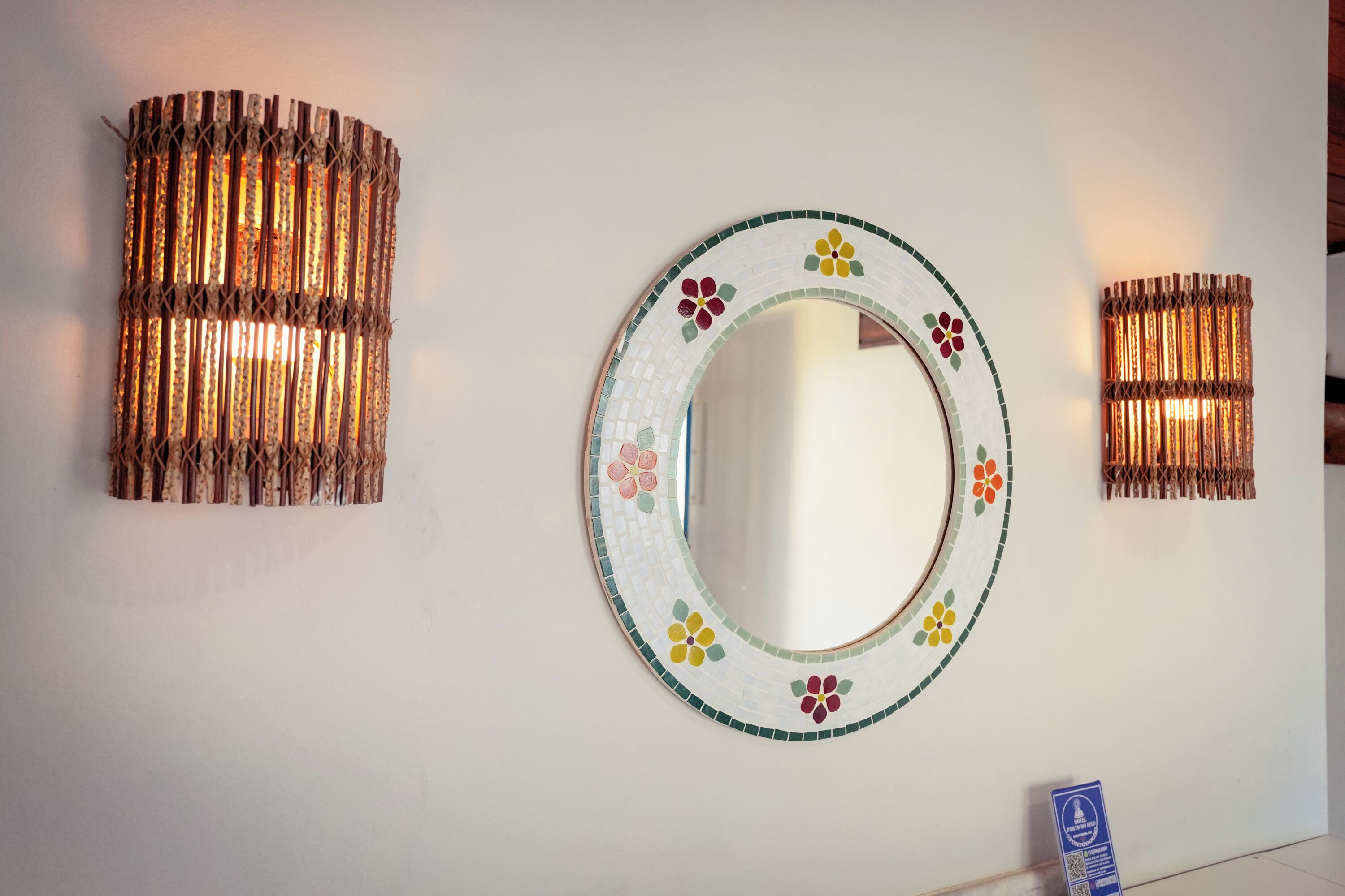 A wall with a round mirror surrounded by a floral-patterned border, flanked by two cylindrical wall sconces with a woven design.