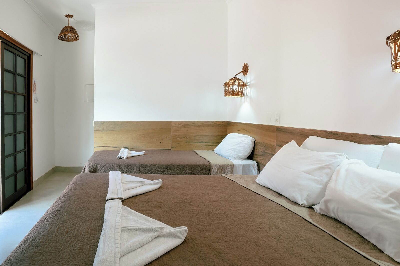 Habitación de hotel con dos camas, cabecero de madera, mesita de noche pequeña y lámpara de pared.