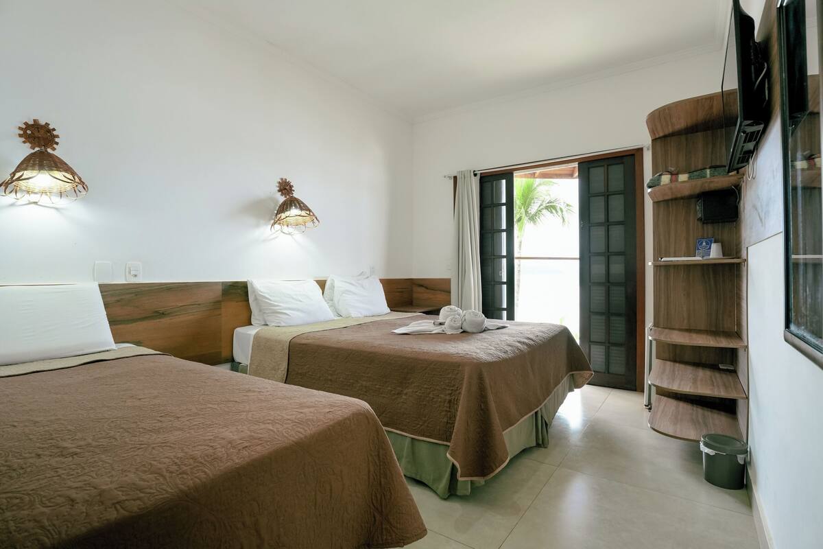 Habitación de hotel con dos camas, un televisor, una estantería de madera y una ventana con vistas a palmeras.