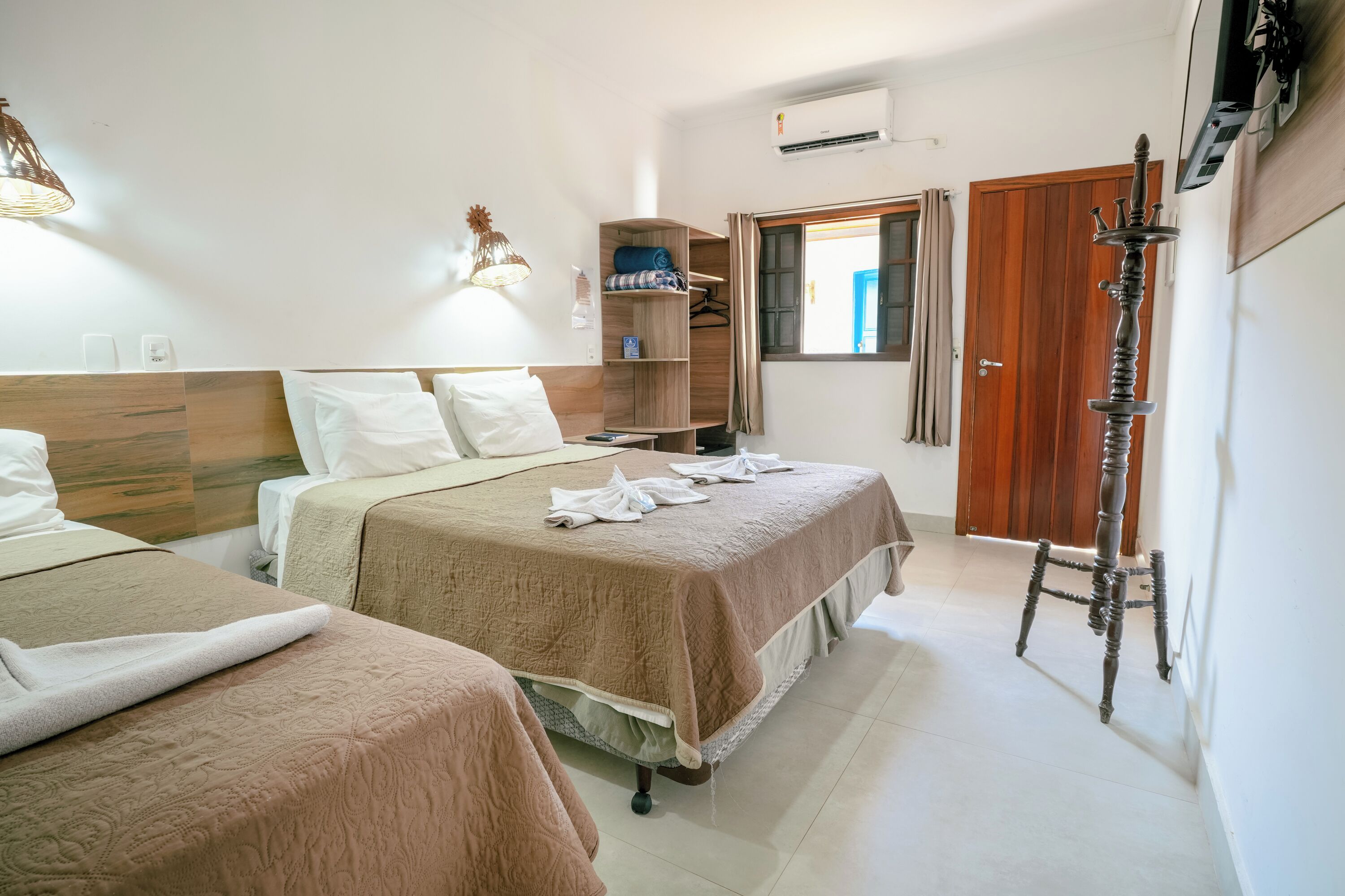 A hotel room with two beds, a wardrobe, a shelf with folded clothes, a standing lamp, and a door.