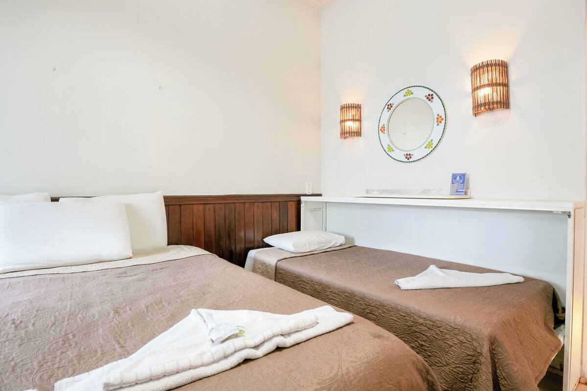 Habitación de hotel con dos camas, cada una con cubrecama marrón y almohadas blancas. Se observa un cabecero de madera, un espejo adosado a la pared con un marco decorativo y dos apliques.