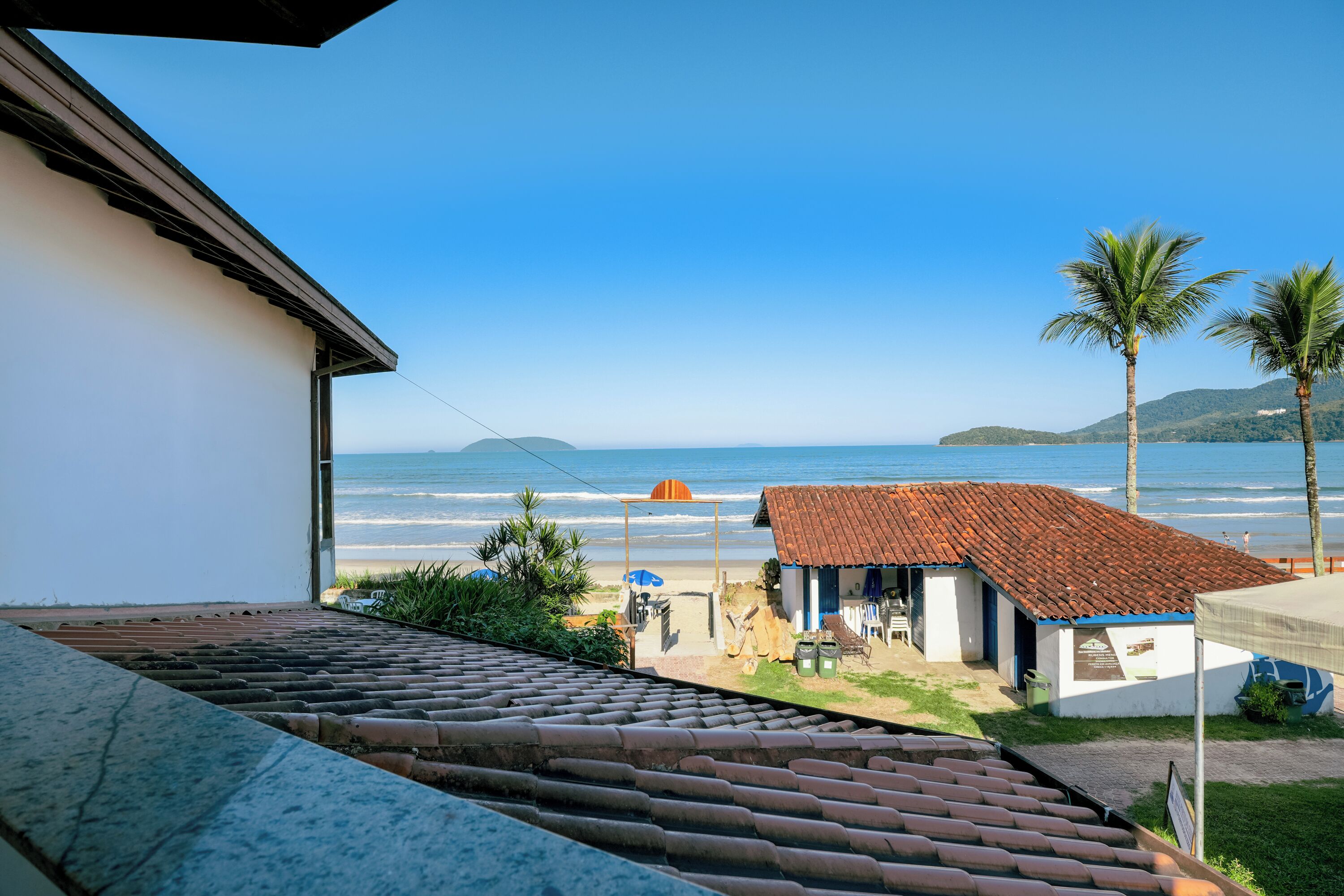 Vista costera desde un balcón con vista a una playa, un edificio pequeño con techo de tejas y palmeras.