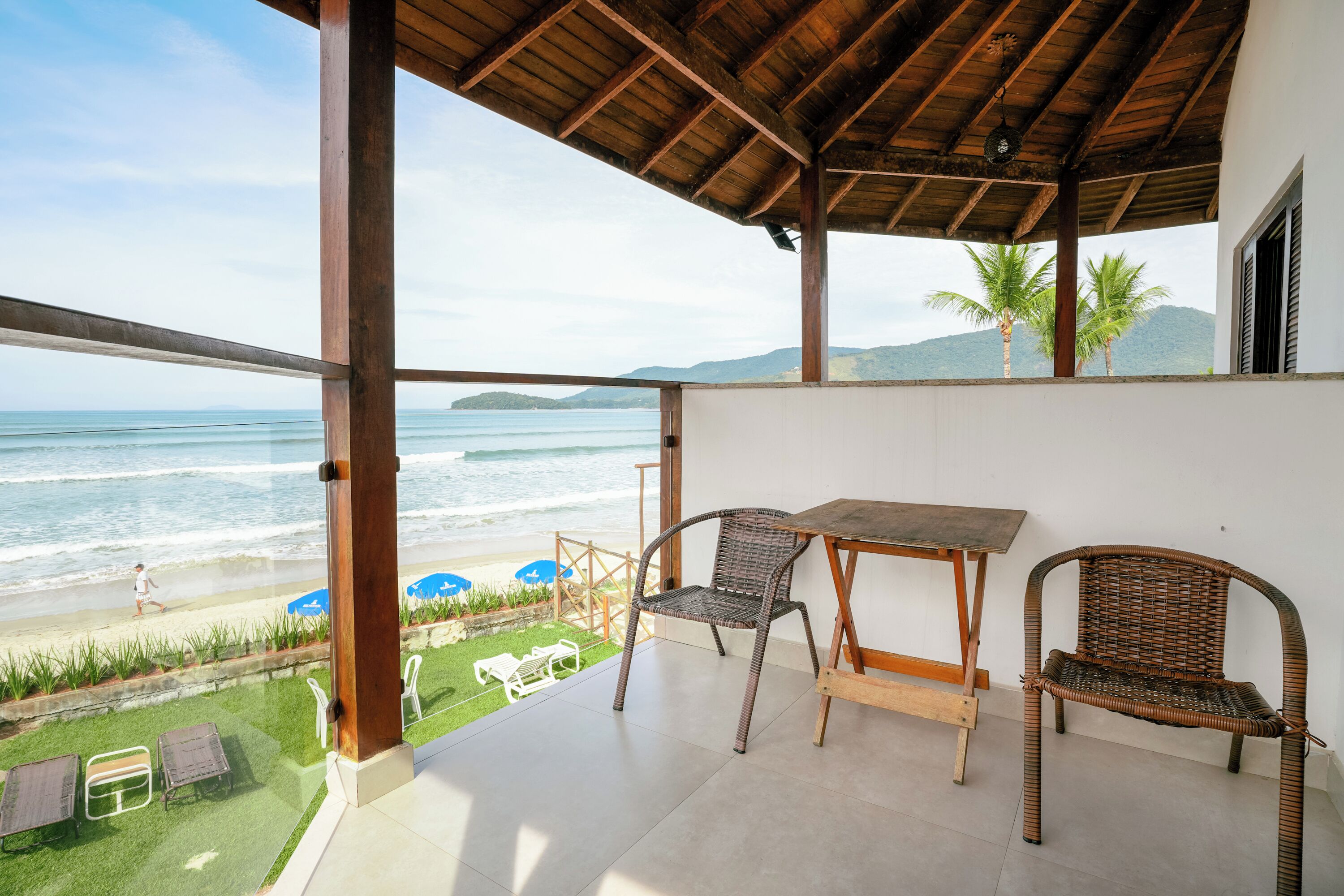 Un balcón con vista al océano, una mesa, dos sillas de mimbre y un área verde con una silla blanca y una mesa pequeña.