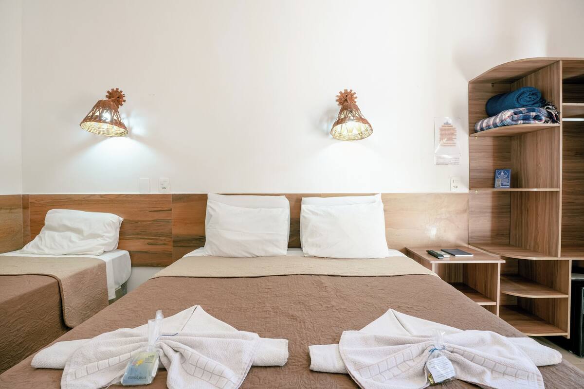 Habitación de hotel con dos camas, cabecero de madera, estante empotrado con toallas dobladas y lámparas fijadas a la pared.