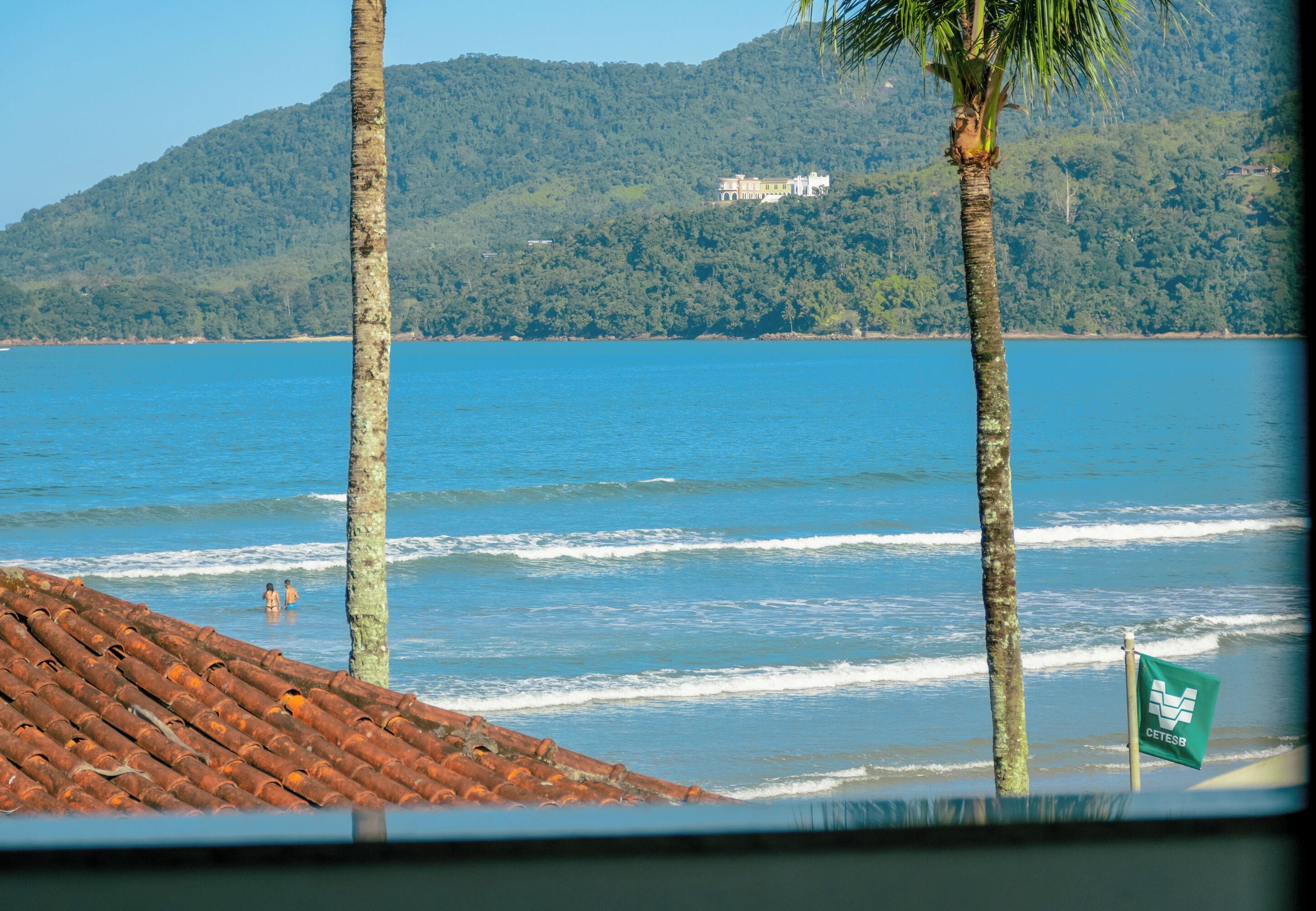 A coastal view with a beach, palm trees, and a building on a hill.