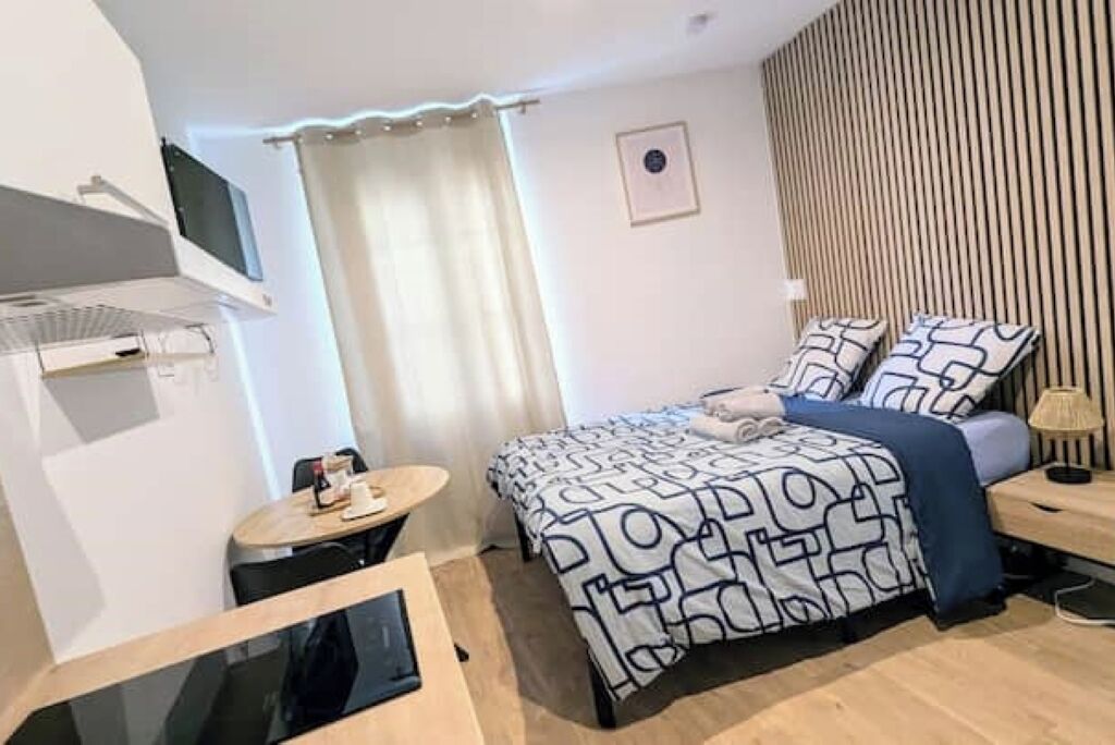 A compact hotel room with a bed, a small desk, a chair, a TV, and a curtain.