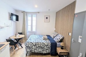 Ein kompaktes Hotelzimmer mit Bett, kleinem Schreibtisch, Stuhl, Wand-Fernseher und einem mit Vorhängen versehenen Fenster.
