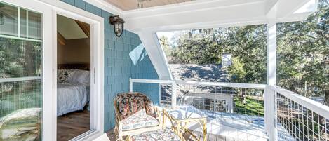 Un balcon extérieur abrité, doté de chaises en osier et d’un lit visible à travers une porte vitrée.