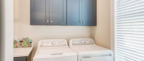 A laundry room with two white front-loading washing machines, a dark blue cabinet above, and a window with blinds.