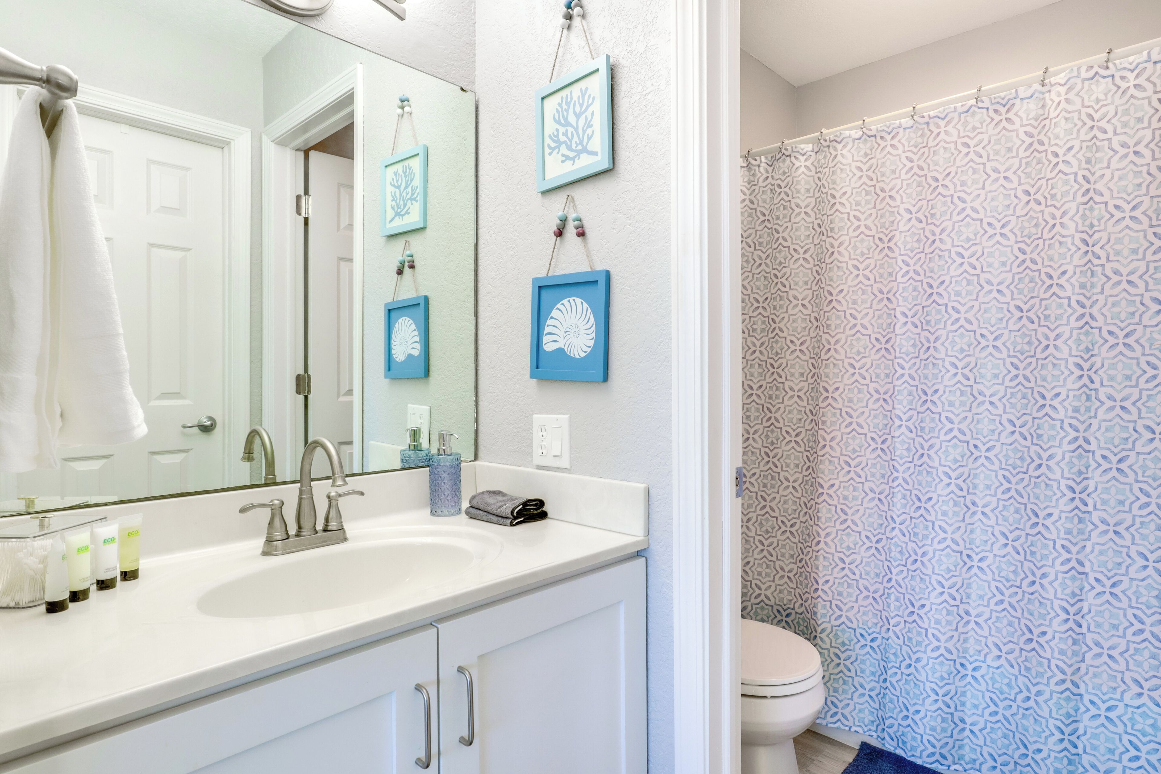 Un bagno con lavandino bianco, specchio e water. Alle pareti sono presenti quadri incorniciati e una tenda da doccia con motivo geometrico.