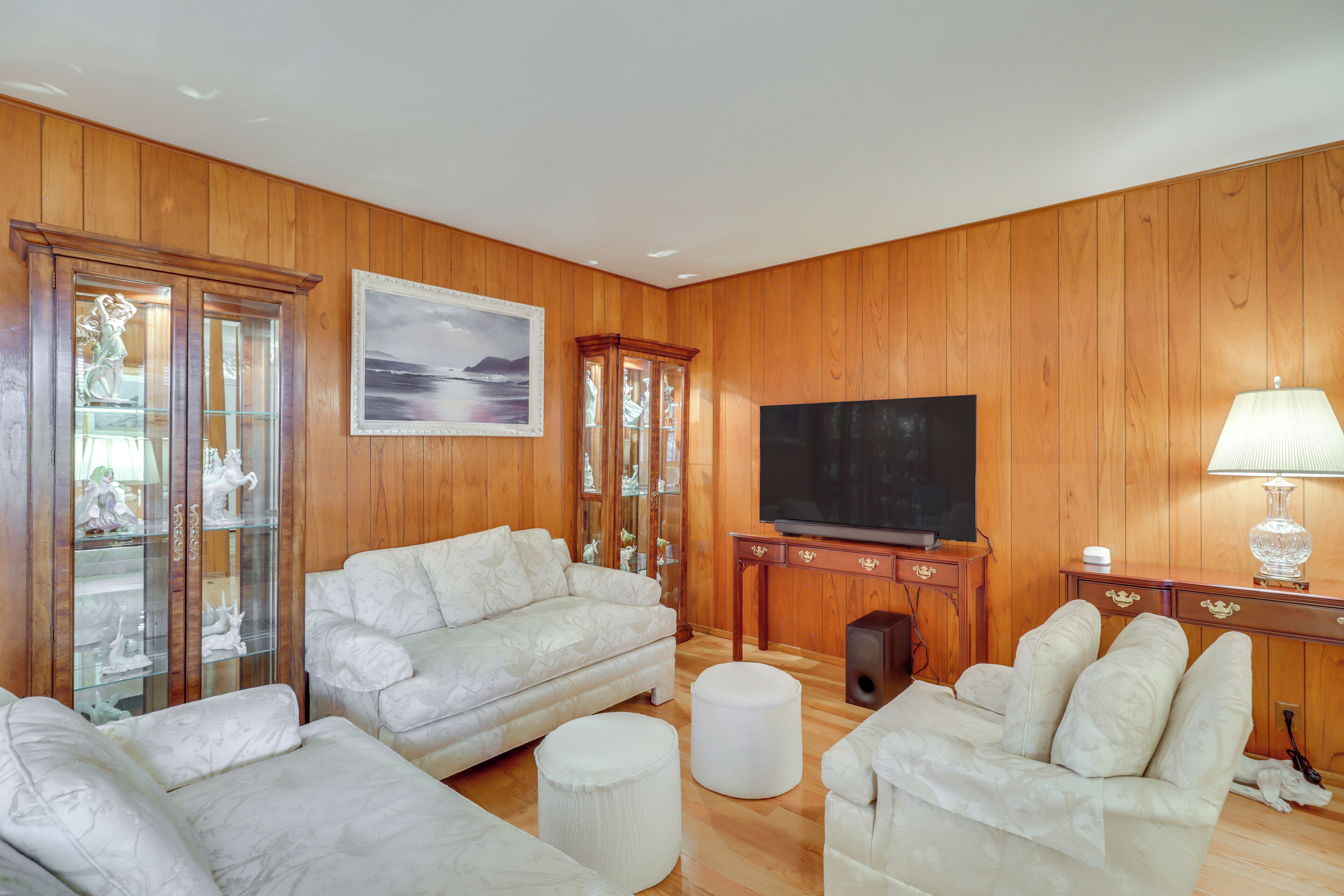 A living room with wooden paneled walls, a large flat-screen TV, a white sofa, a small ottoman, and a glass cabinet with figurines.