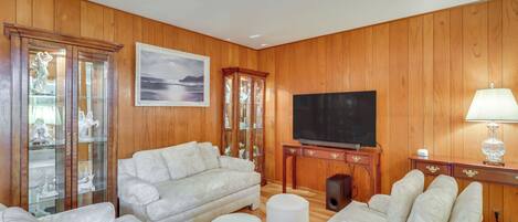 A living room with wooden paneled walls, a large flat-screen TV, a white sofa, a small ottoman, and a glass cabinet with figurines.