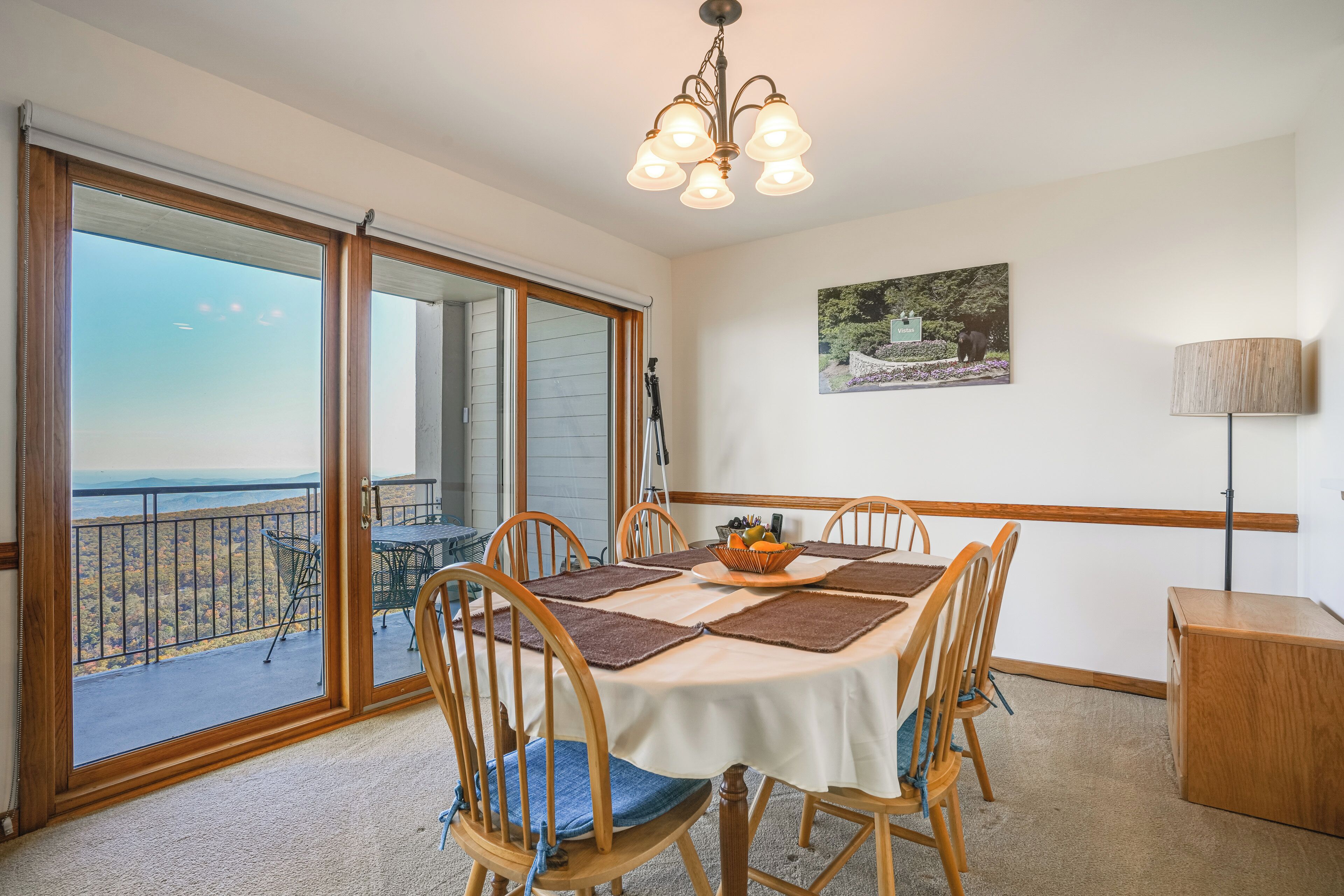 A dining area with a table set for four, wooden chairs, a chandelier, a painting on the wall, and a view through the glass doors.