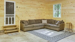 A wooden cabin interior with a L-shaped sectional sofa, a welcome mat, and a window with a view of trees. - Golf Getaway in Cadillac: Deck + Putting Green (Cadillac)