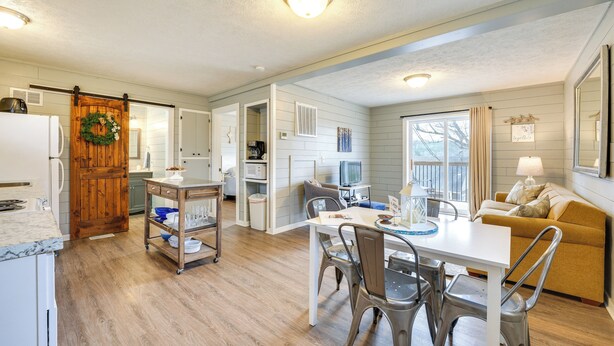 A modern kitchen with a wooden door, a dining area with a white table and metal chairs, and a living room with a yellow sofa and a TV. - Walk to Lake & Trails: Cape Fair Retreat w/ Grill! (Cape Fair)