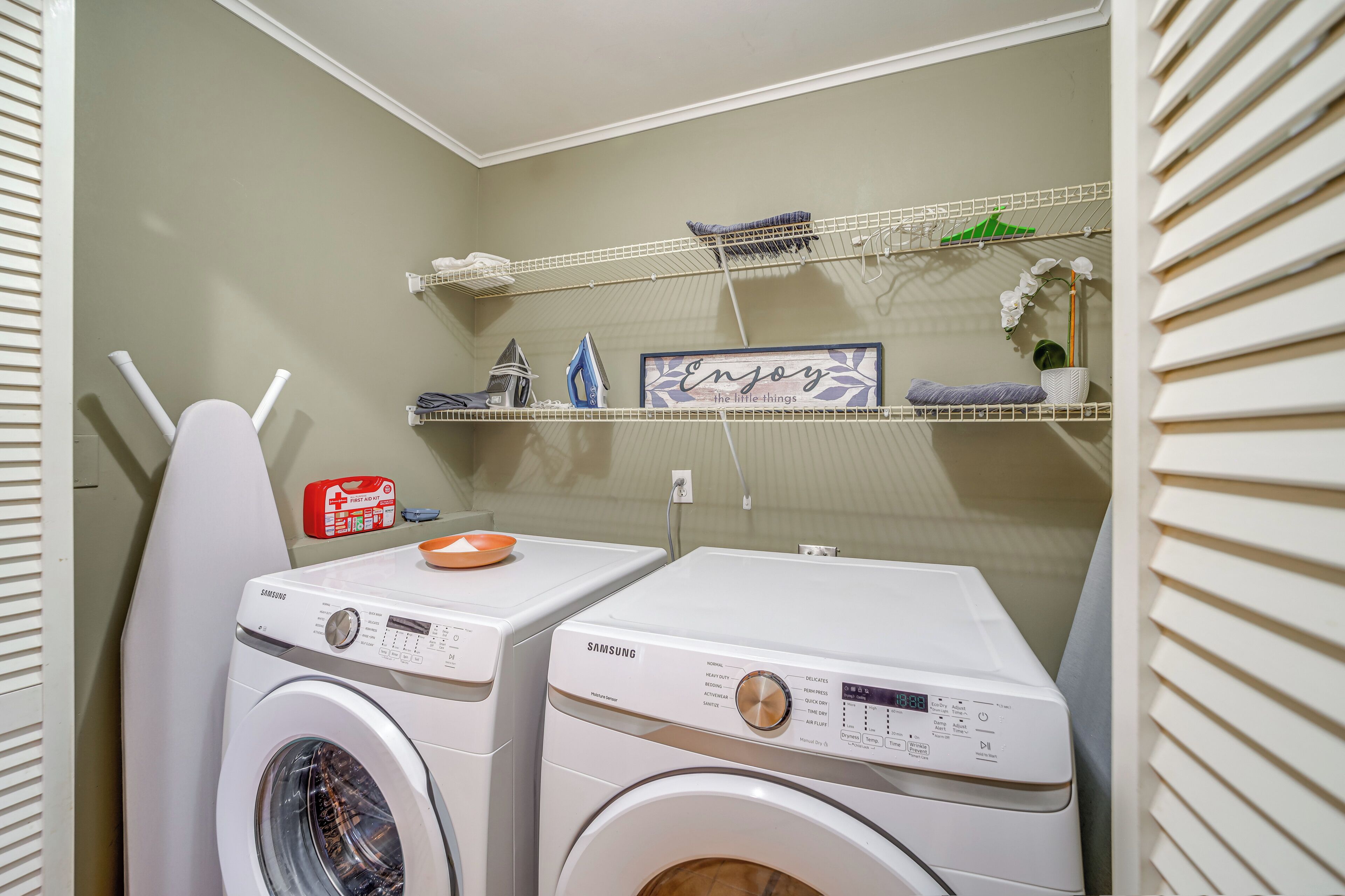 A laundry area with two white Samsung washing machines, a shelf with various items, and a sign that says "Enjoy the little things."