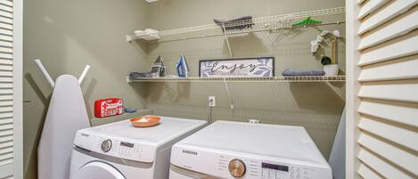 A laundry area with two white Samsung washing machines, a shelf with various items, and a sign that says "Enjoy the little things."