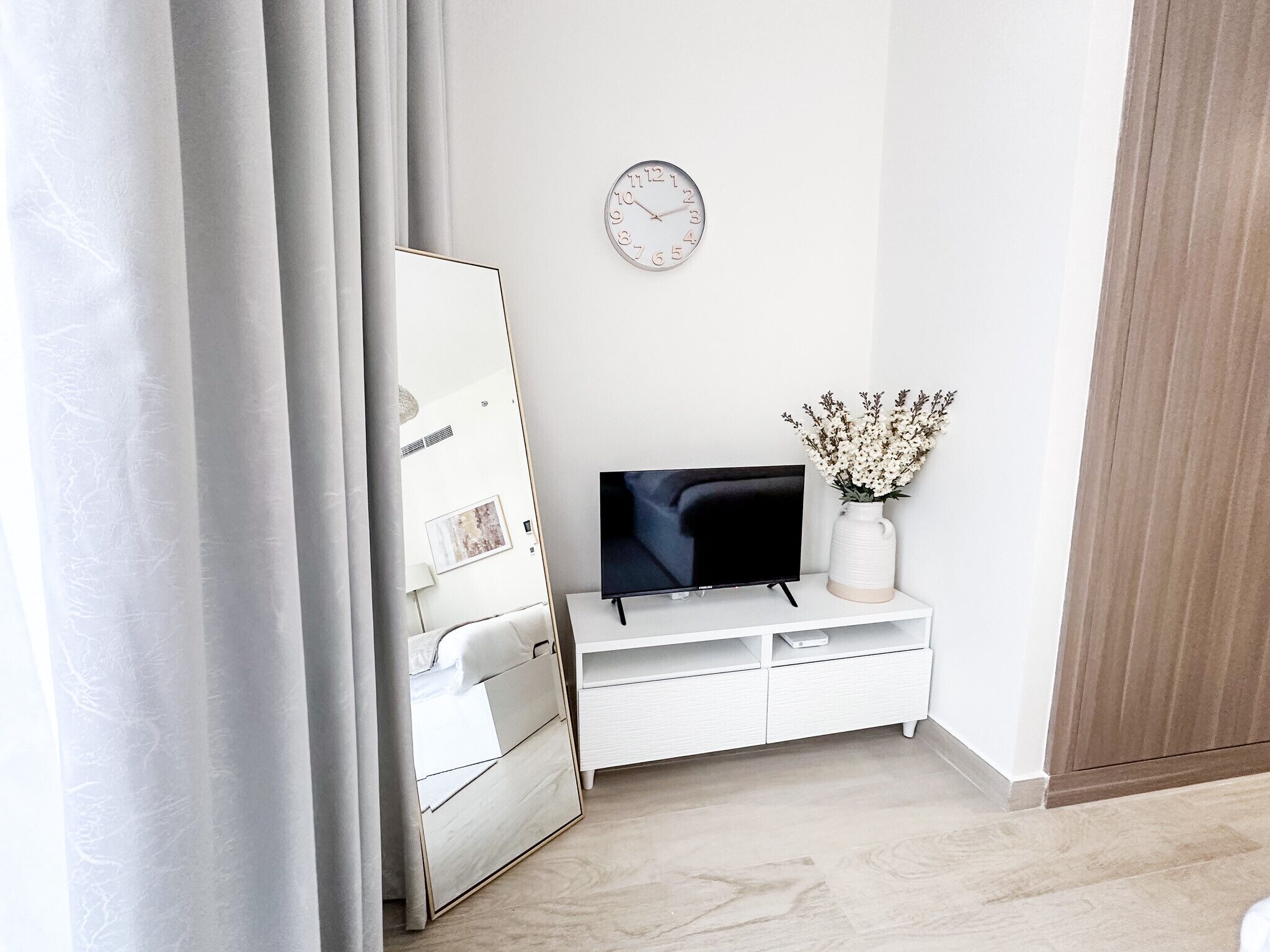 A modern room with a flat-screen TV on a white console, a clock on the wall, and a vase with flowers on a shelf.