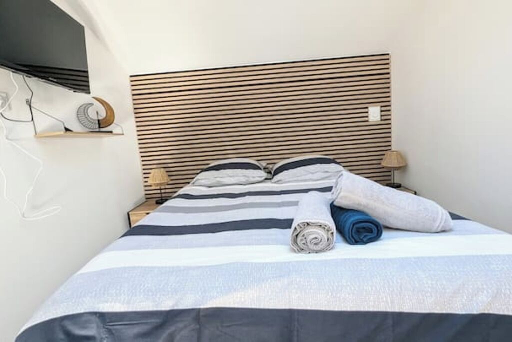 Een net opgemaakt bed met strepen op het beddengoed, een houten hoofdbord en een opgerolde handdoek aan de zijkant.