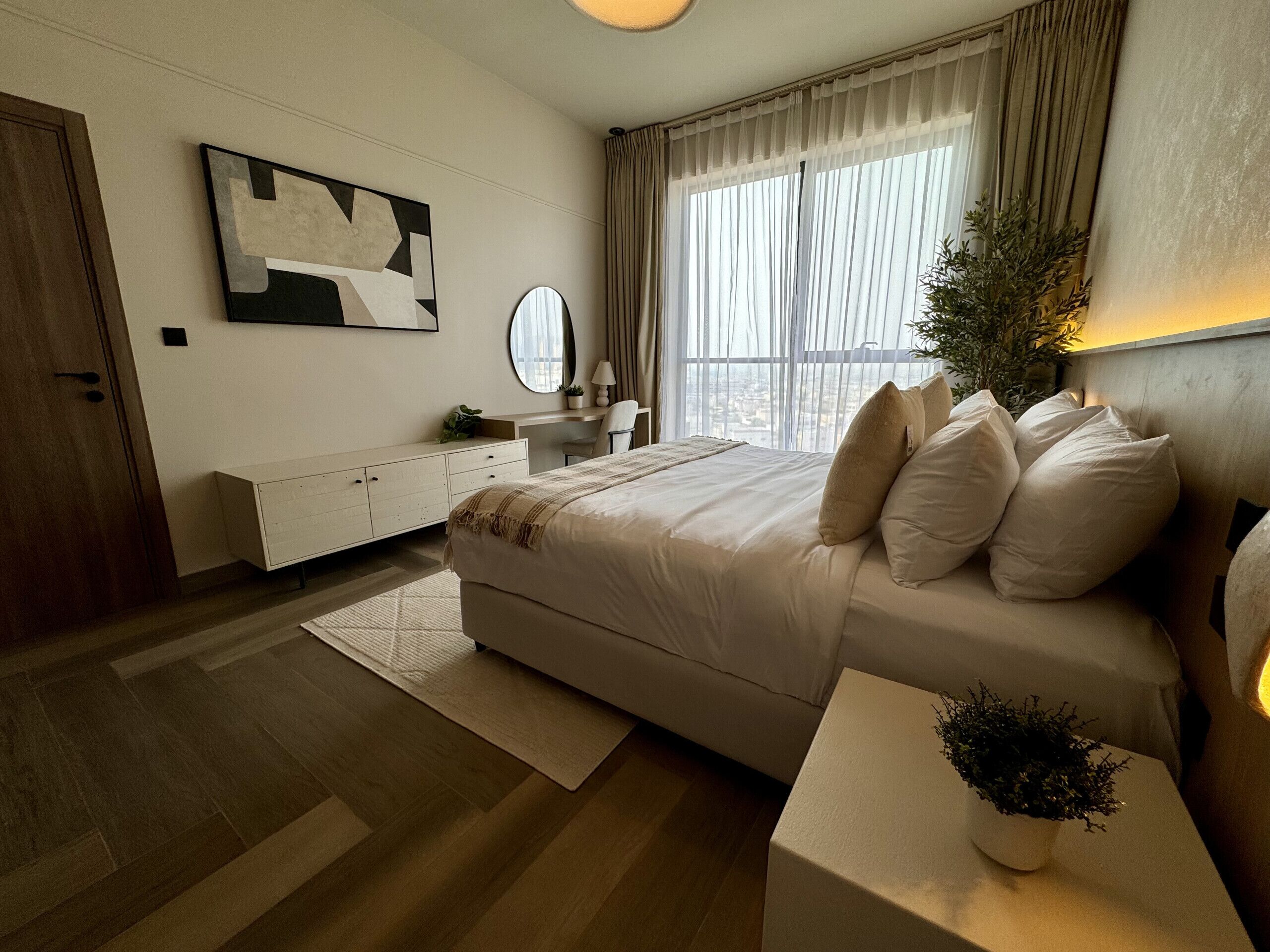 Un dormitorio moderno con una cama grande, una mesita de noche con una planta, un aparador, un espejo y una ventana con cortinas.