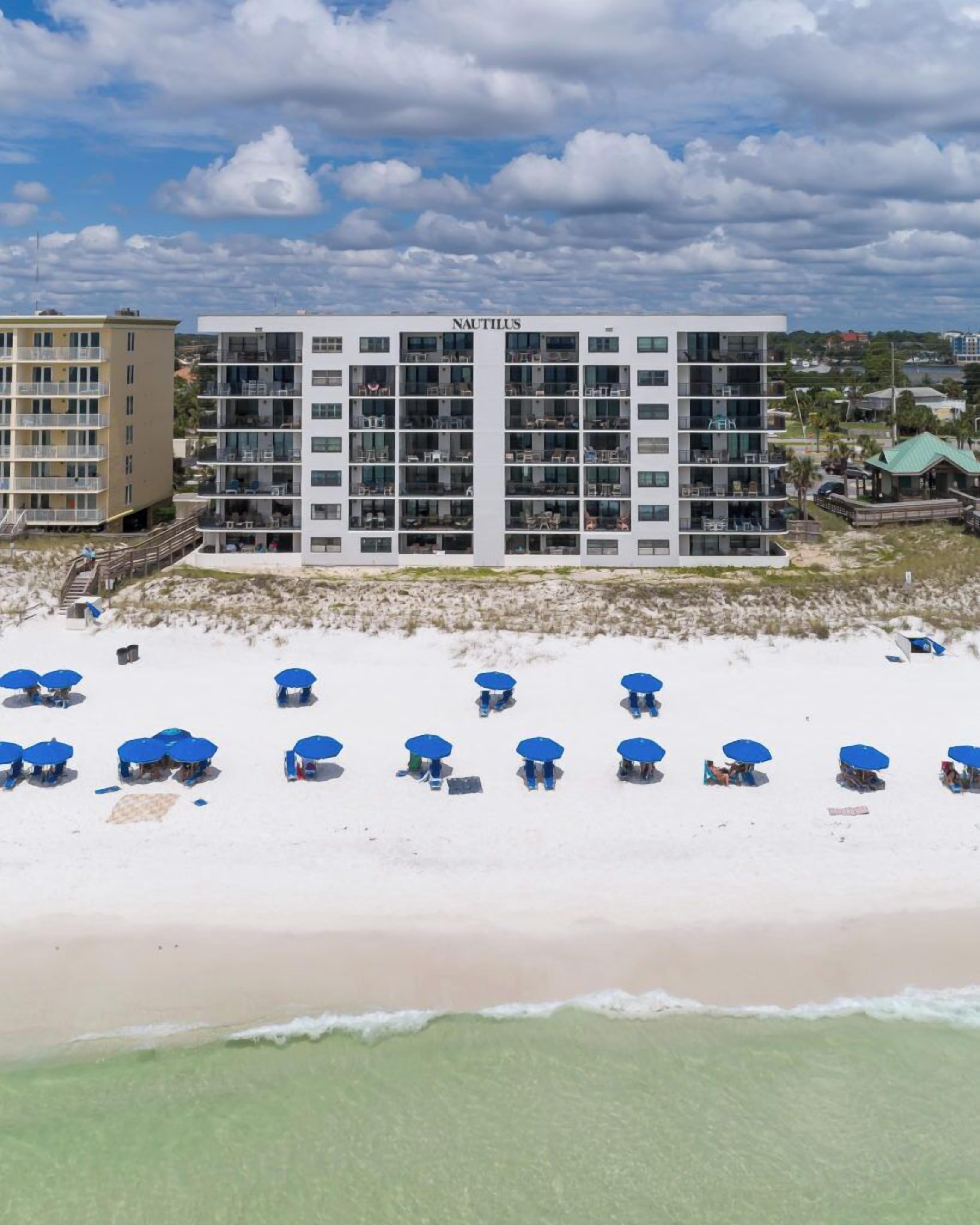 Een strandhotel met meerdere balkons, een zandstrand met blauwe parasols en een heldere lucht.