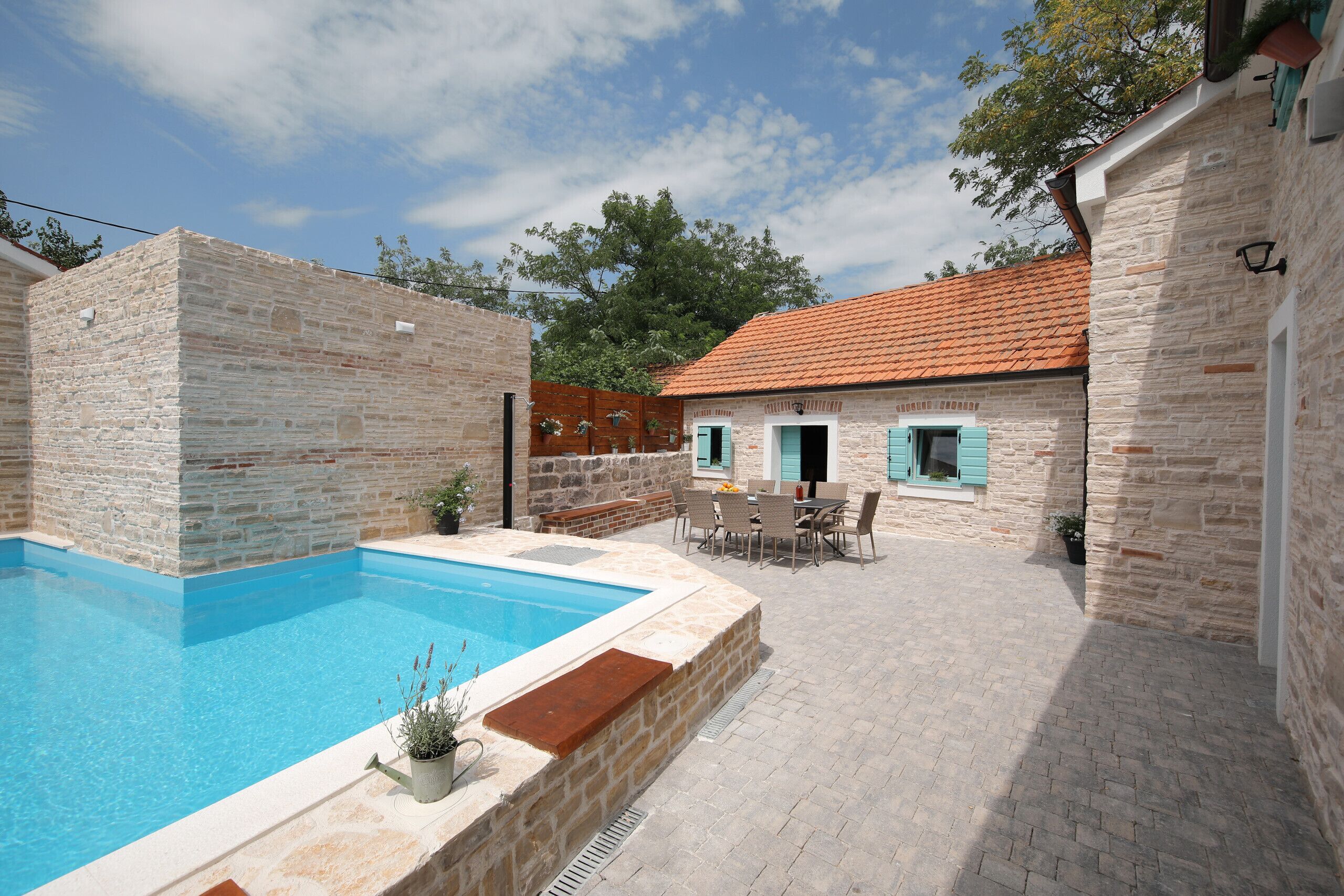 A swimming pool with stone walls, a patio with chairs, and a building with a tiled roof.