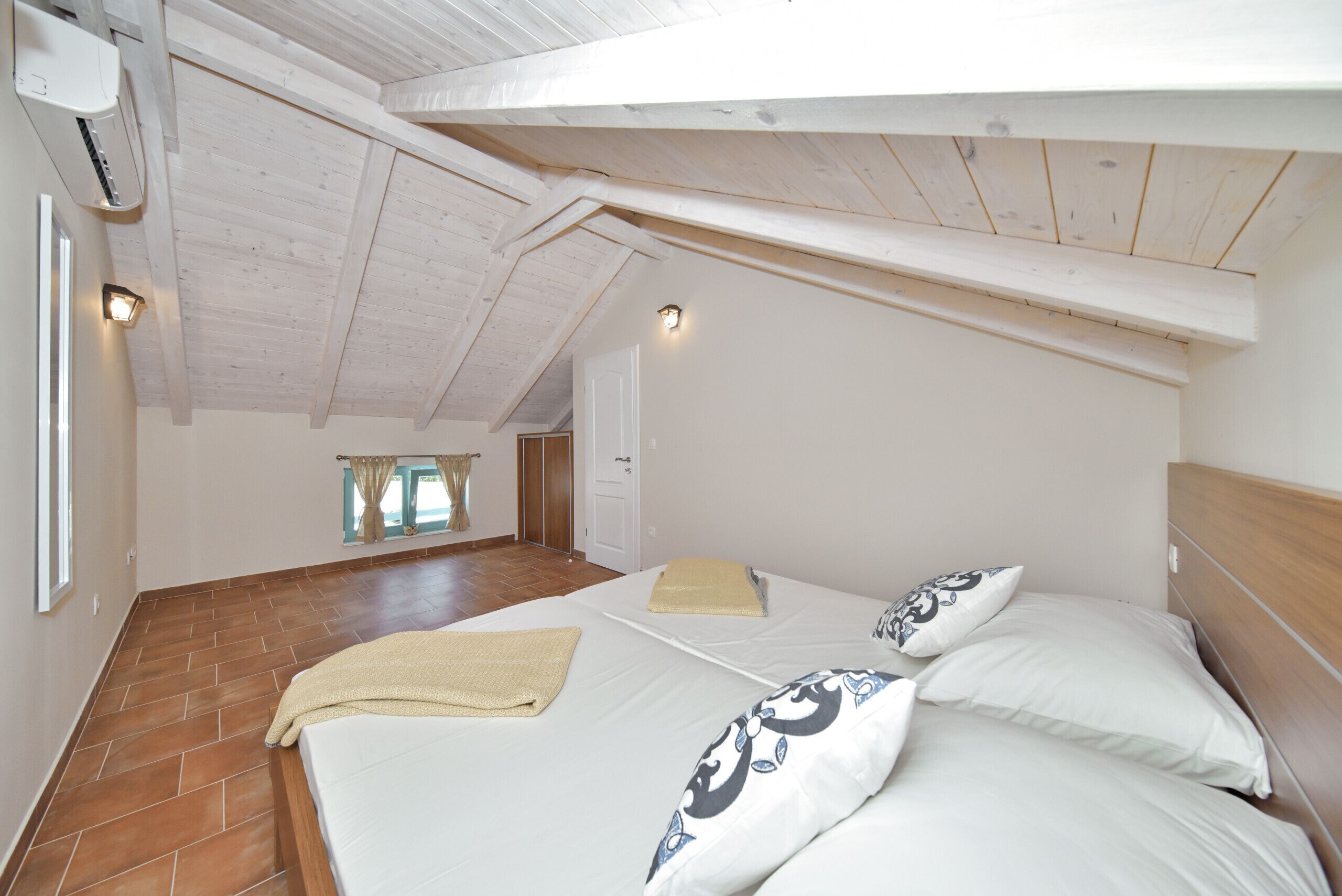 Una camera da letto con soffitto a falde, letto con lenzuola bianche, poggiatesta in legno e finestra con tende.